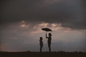 "The Right Thing" - a photo of an adult holding an umbrella, standing in front of a child. Background includes dark clouds.